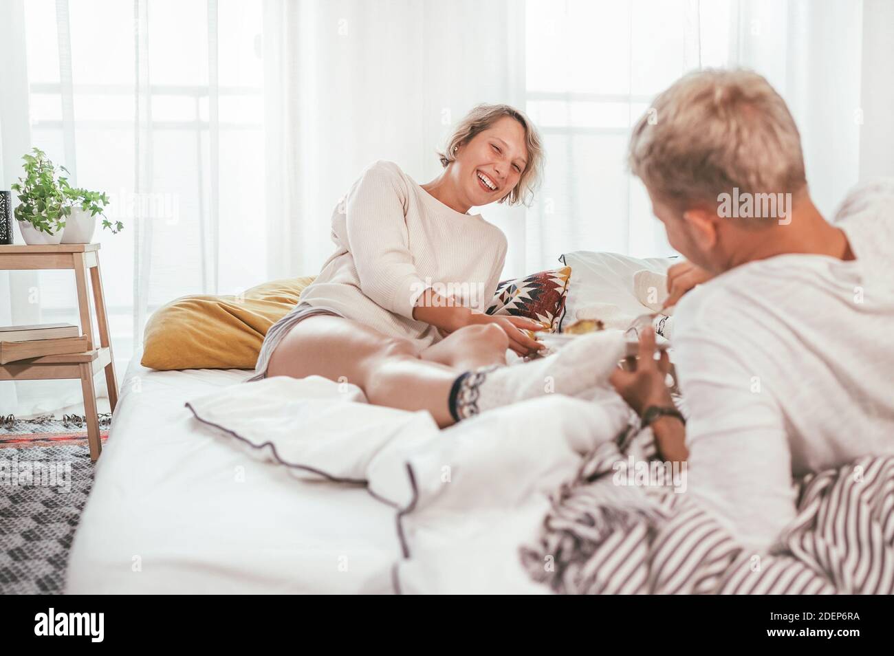 Lachend blonde Haare junge Erwachsene Paar in Pyjamas faul entspannen in einem gemütlichen Bett im Schlafzimmer liegen und mit einem friedlich unbeschwerten Plaudern. Paar Stockfoto