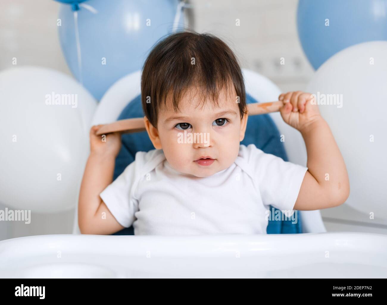 Kleiner Junge, der zu Hause in einem blauen Hochstuhl sitzt Auf der weißen Küche und spielen mit Holzlöffel auf Hintergrund mit Ballons Stockfoto