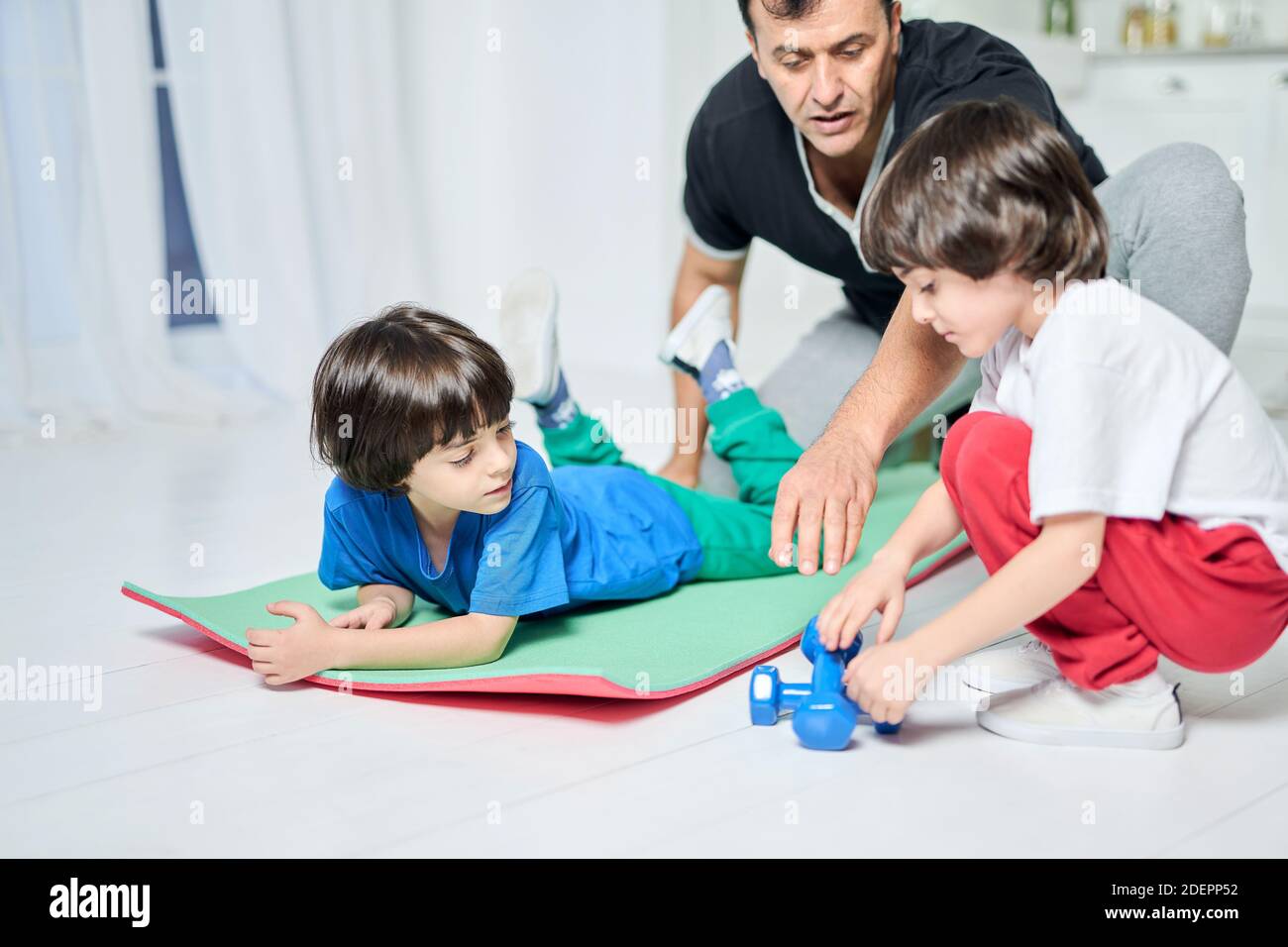 Fürsorglicher hispanischer Vater, der Zeit mit seinen zwei kleinen Jungen verbringt, zusammen trainiert, während er auf einer Matte im häuslichen Interieur sitzt. Vaterschaft, Sport, Bildungskonzept Stockfoto