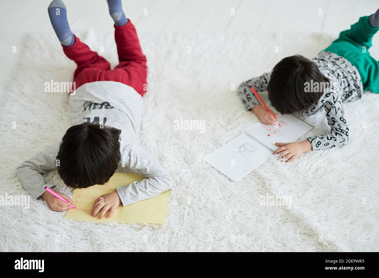 Zwei kleine lateinische Jungen, Zwillingsbrüder zeichnen Bilder mit bunten Bleistiften in Papier-Album, während sie auf dem Boden liegen. Geschwister in kreative Tätigkeit zusammen beteiligt. Fernunterricht für Kinder Stockfoto