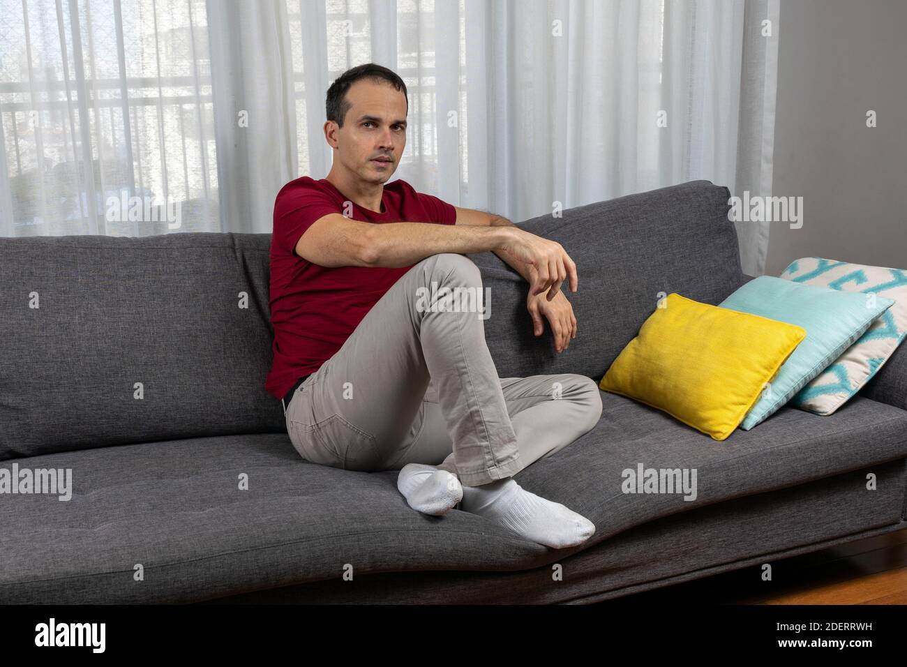 Reifer Mann (44 Jahre) sitzt auf der Couch, mit Socken und neben bunten Kissen. Stockfoto