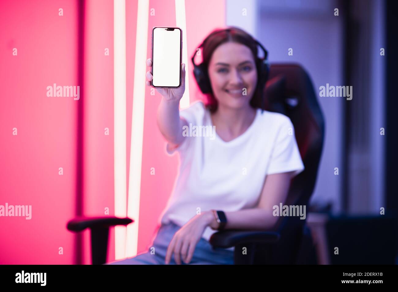 Lächelndes Gamer Mädchen trägt Headset zeigt Smartphone und zeigt auf leeren Bildschirm mit ihrem Finger. Stockfoto