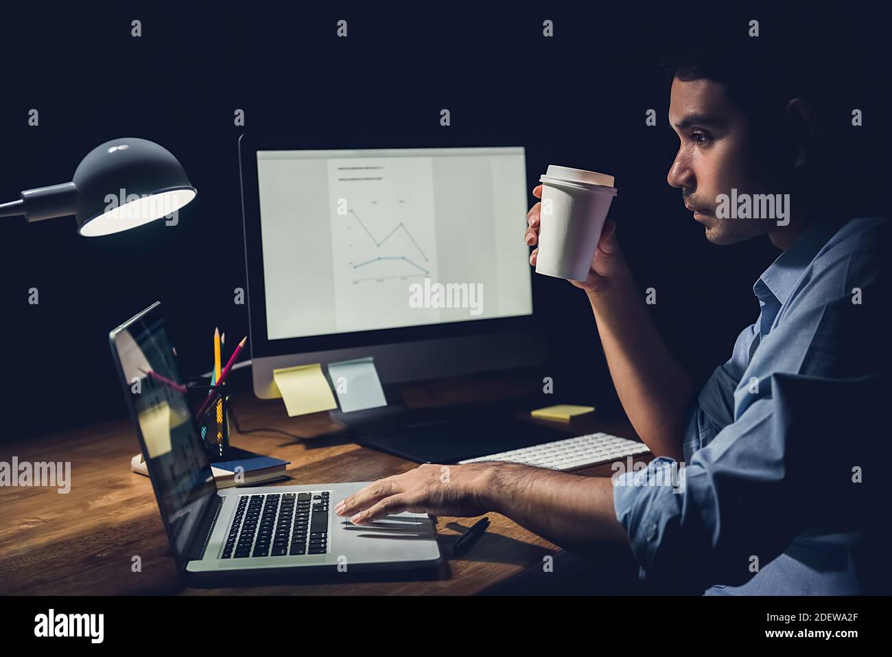Schläfriger, müde Geschäftsmann, der spät in der Nacht Überstunden im Hotel verbracht hat Büro hält Kaffeetasse und konzentriert sich auf die Arbeit mit Laptop Computer an seinem Schreibtisch Stockfoto