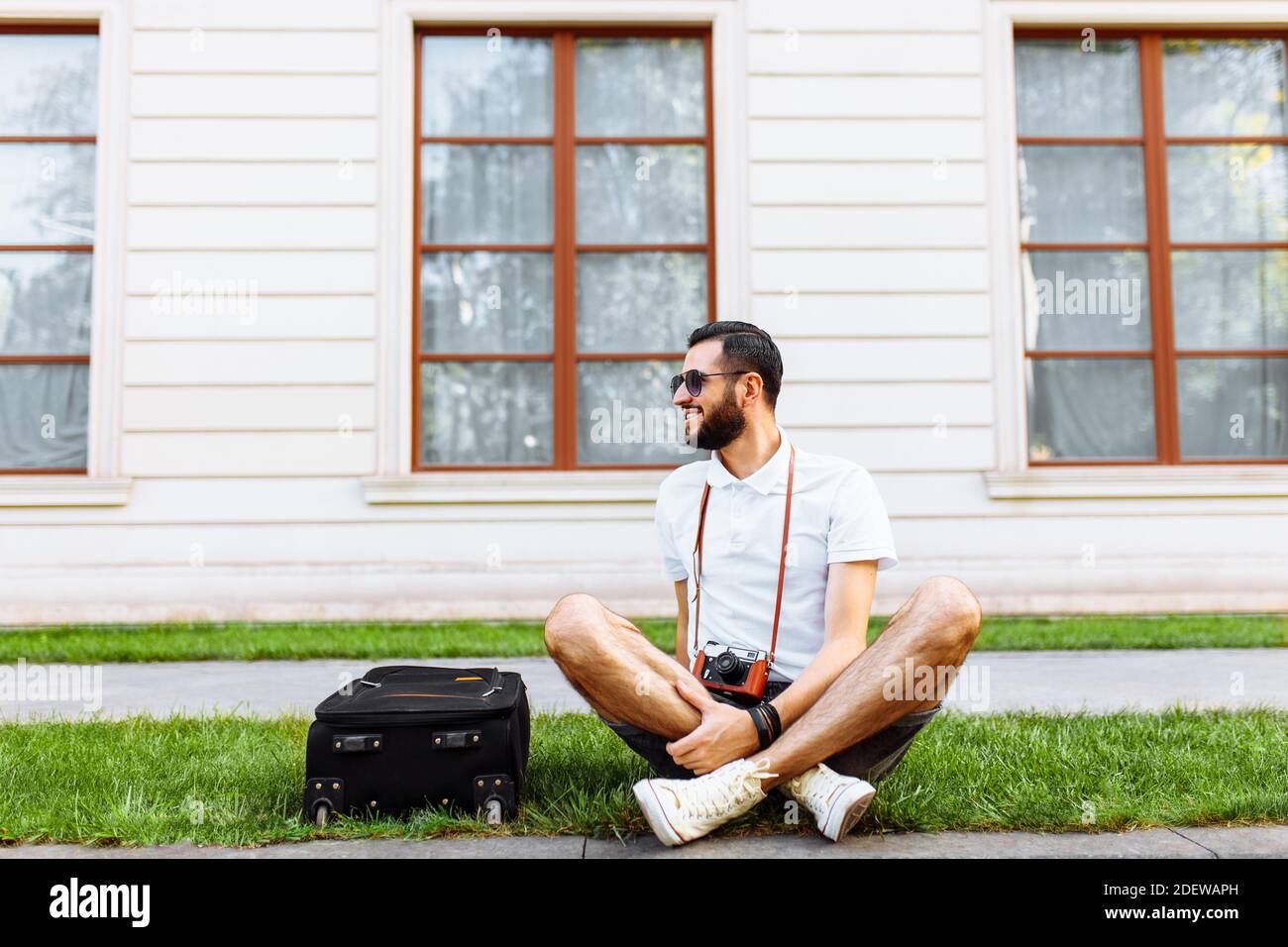Stilvoller Hipster mit Koffer und Kamera, ein Reisender sitzt auf der Wiese im Park Stockfoto