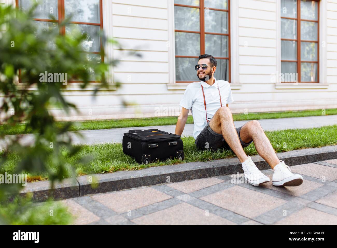 Stilvoller Hipster mit Koffer und Kamera, ein Reisender sitzt auf der Wiese im Park Stockfoto