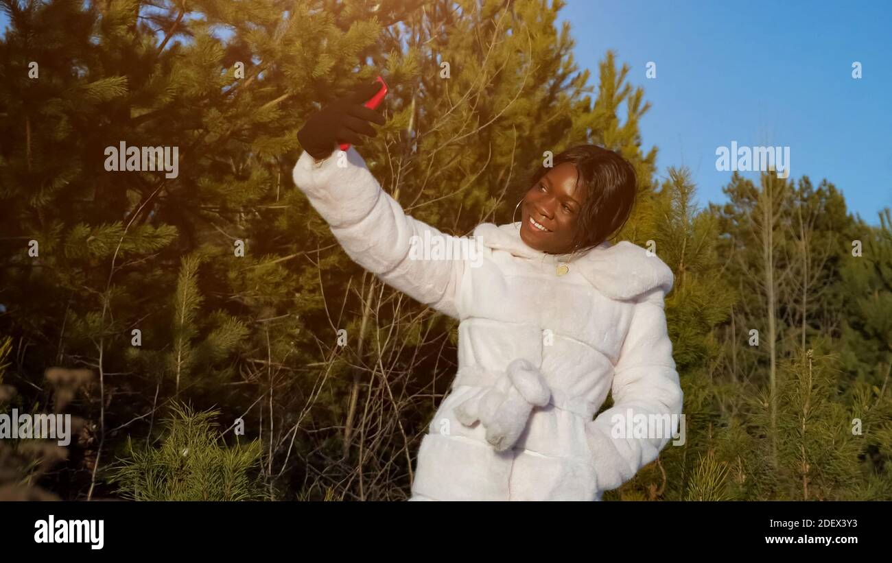 Schöne schwarze Frau in Pelzmantel macht Selfie mit Smartphone gegen wilden Winter immergrünen Wald unter klarem blauen Himmel Stockfoto