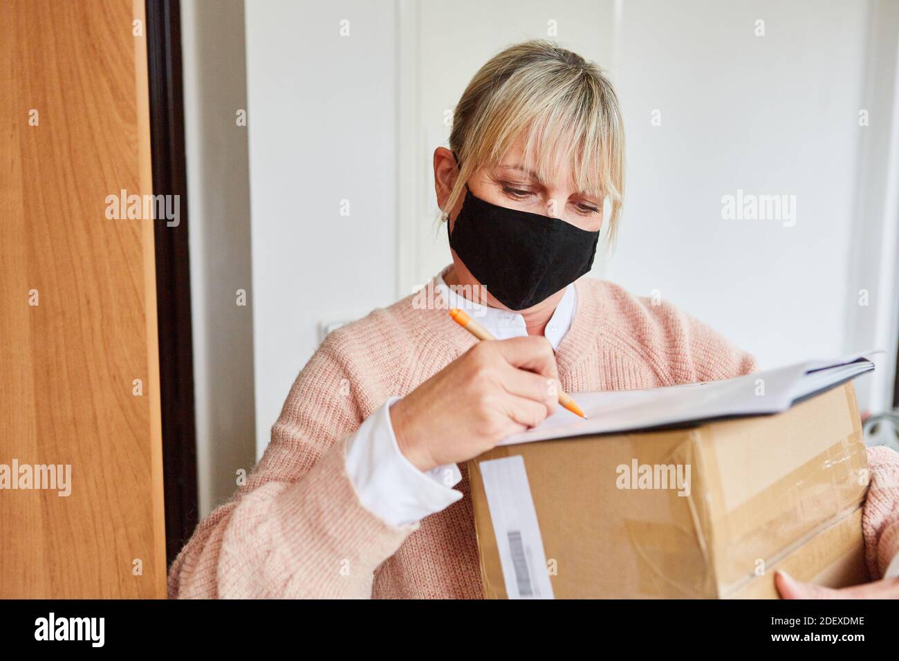 Frau mit Gesichtsmaske vor der Wohnungstür Gibt dem Zustelldienst eine Unterschrift Stockfoto