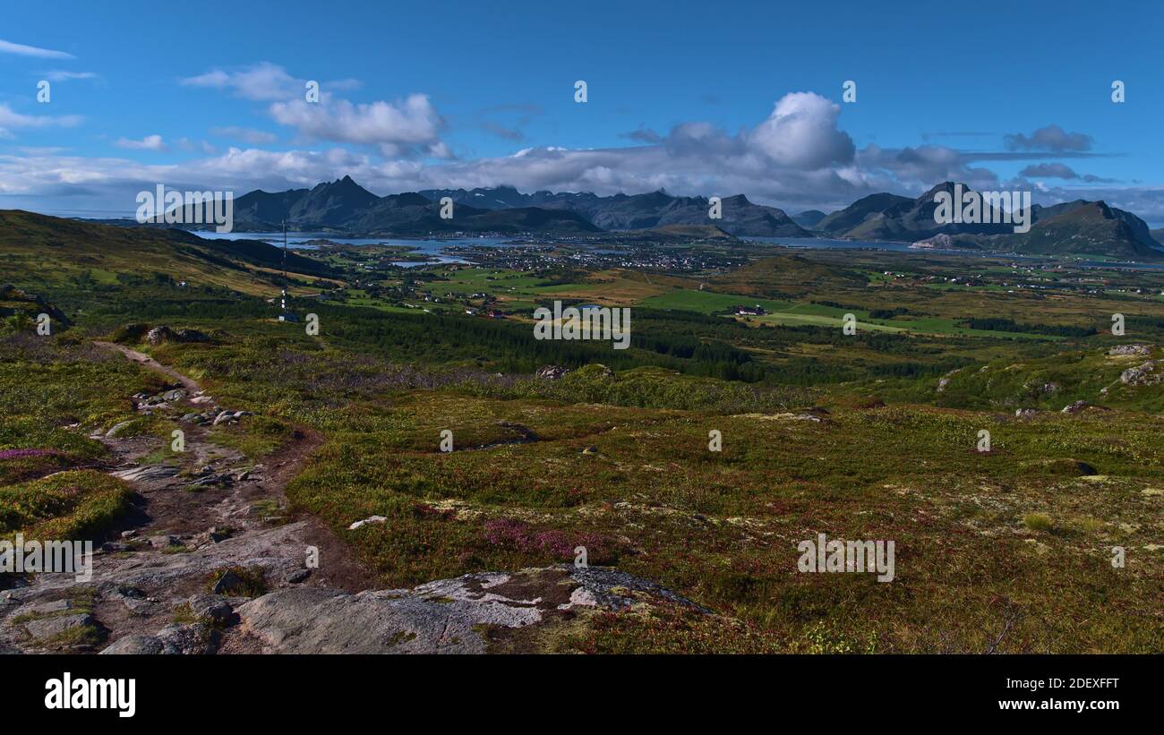 Schöner Panoramablick über den Westen der Insel Vestvågøy mit felsigen Wanderweg, grüner Vegetation, Antennenturm, Leknes Stadt und schroffen Bergen. Stockfoto