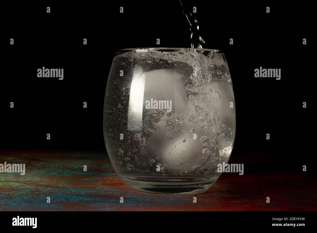 Ein Glas Eiswasser, das auf dunklem Grund und rustikaler Oberfläche mit sprudelndem Wasser gefüllt ist. Stockfoto