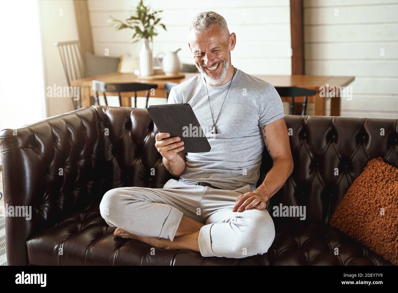 Glücklicher Mann mittleren Alters in legerer Kleidung mit Videoanruf Auf einem digitalen Tablet, während Sie in Lotuspose auf dem Sofa sitzen Im Wohnzimmer Stockfoto
