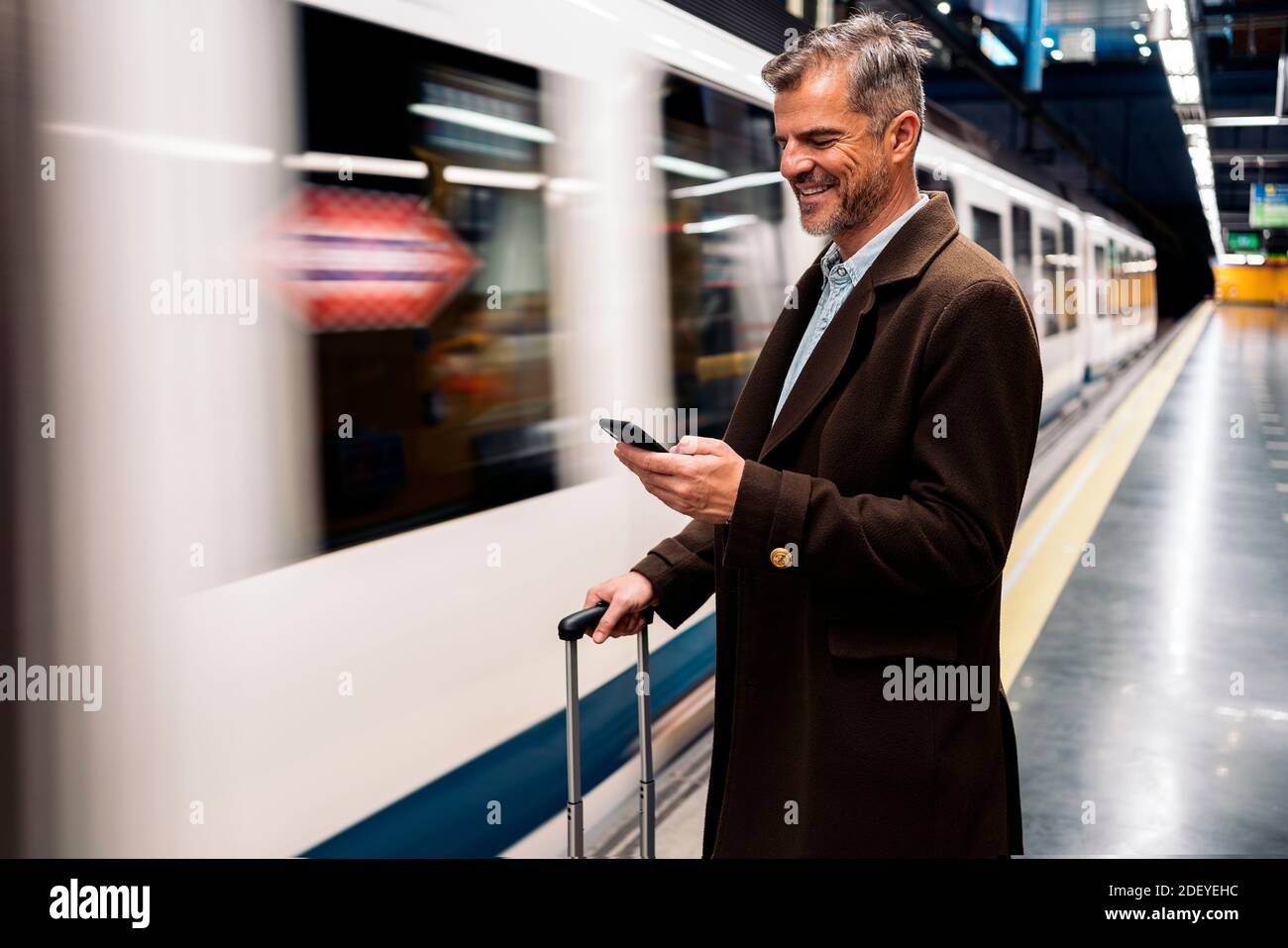 Stock Foto von einer Seitenansicht eines Geschäftsmanns tippen in seinem Smartphone wartet auf die U-Bahn in der Plattform nehmen. Er lächelt und trägt Casua Stockfoto