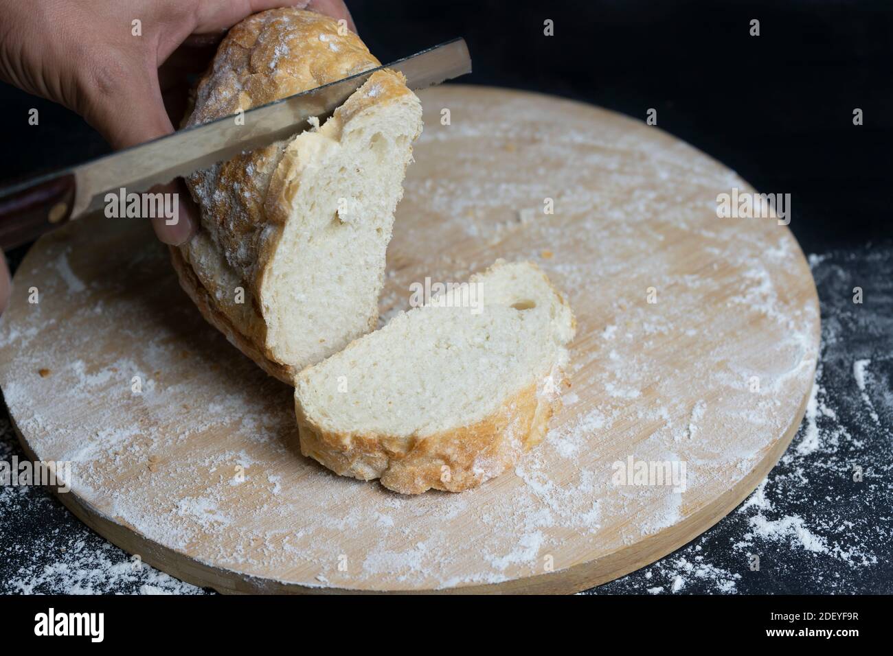 Hand schneiden Weizen traditionelle Brot mit einem Messer auf mehligen Holzbrett.Rustikale Bäckerei Konzept.Dunkler Hintergrund. Stockfoto