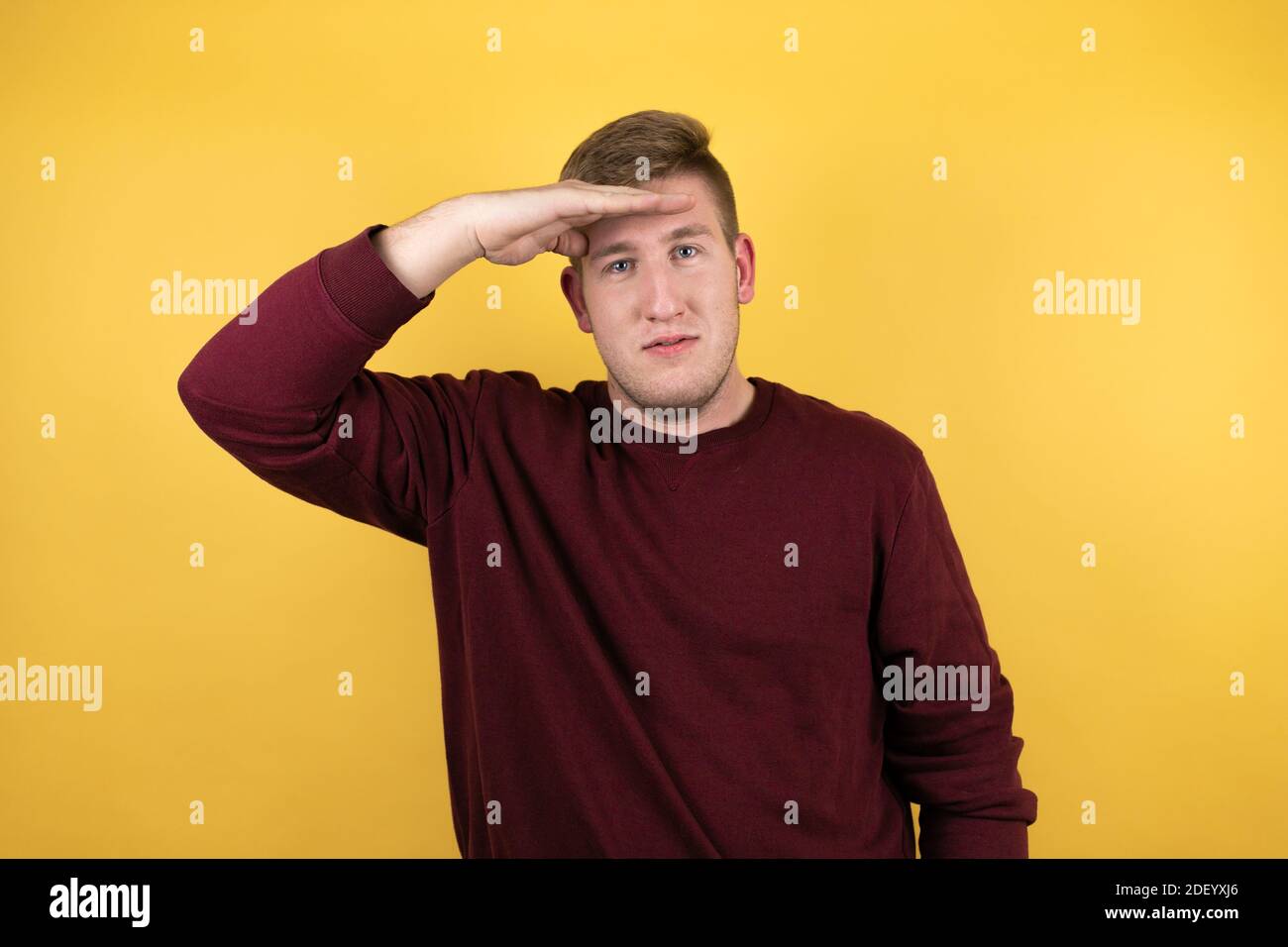 Junge blonde Mann trägt einen lässigen roten Pullover über gelben Hintergrund sehr glücklich und lächelnd suchen weit weg mit Hand über Kopf. Suchkonzept. Stockfoto