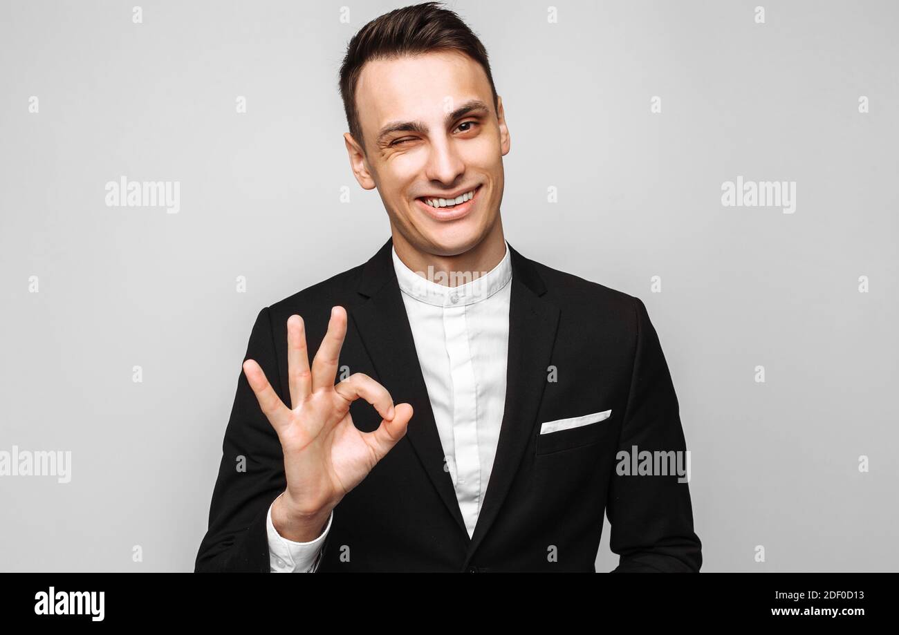 Porträt eines jungen, gutaussehenden glücklichen Mannes, in einem Geschäftsanzug, lächelnd und mit einer OK-Geste, auf grauem Hintergrund. Positive menschliche Emotionen, Gesten Stockfoto