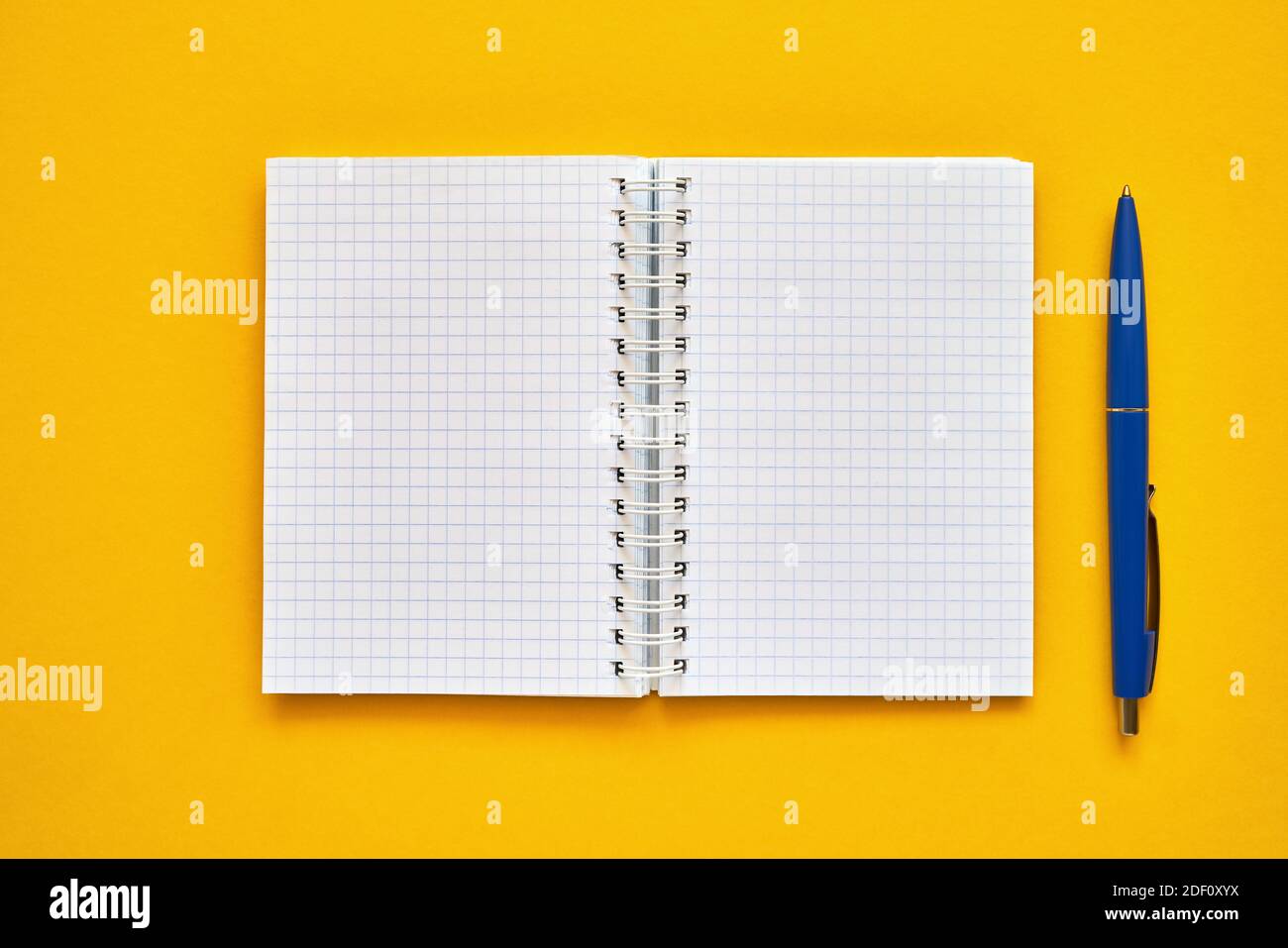 Draufsicht auf ein offenes Notizbuch mit leeren quadratischen Seiten und blauem Stift. Schulnotizbuch auf gelbem Hintergrund, Spiralnotizblock. Zurück zur Schule Konzept. Stockfoto