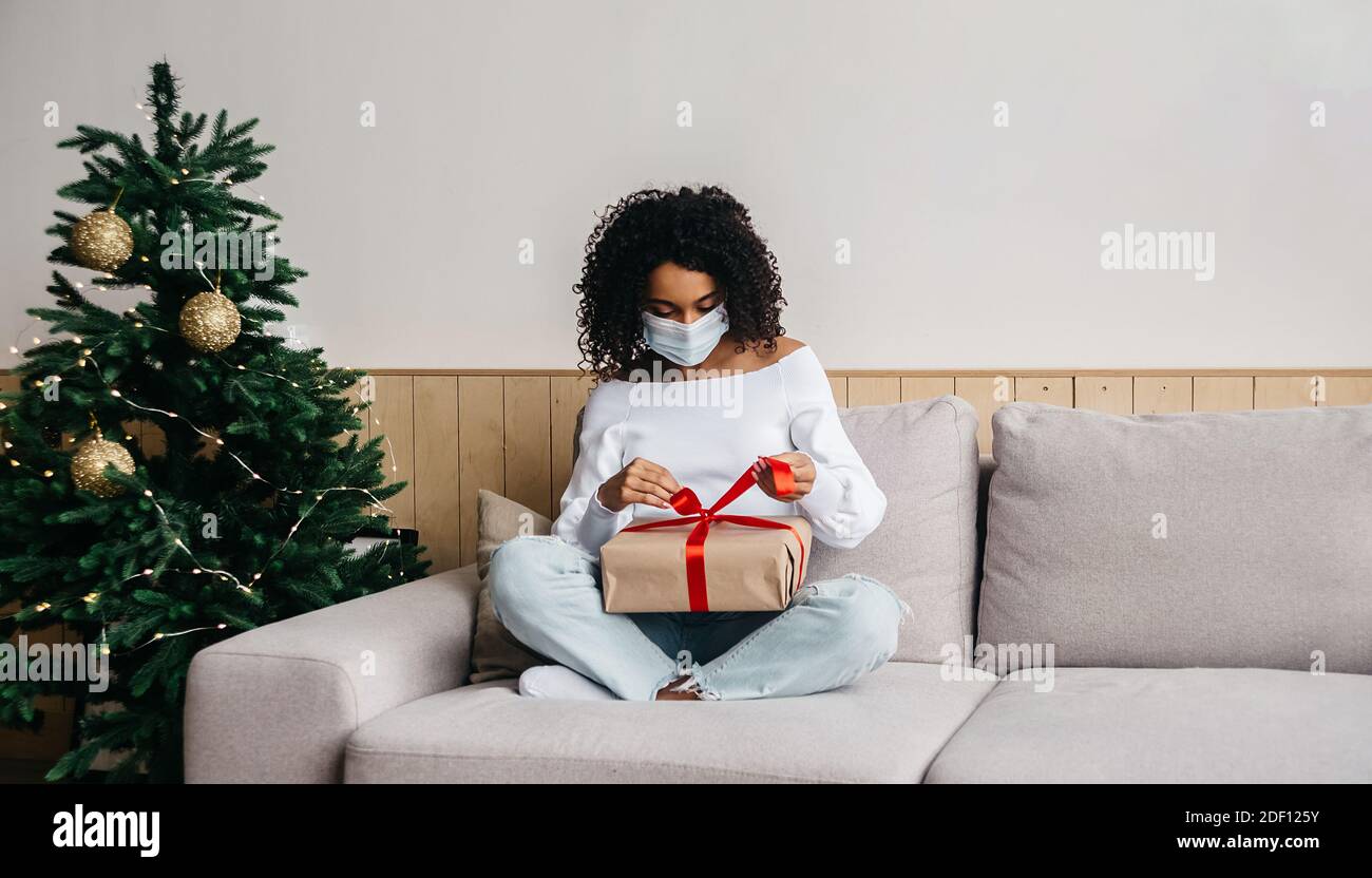 Afrikanische Frau trägt Schutzmaske, packt Geschenke aus. Online-Weihnachtseinkäufe und das Versenden von Geschenken per Post während einer Coronavirus-Pandemie. Stockfoto