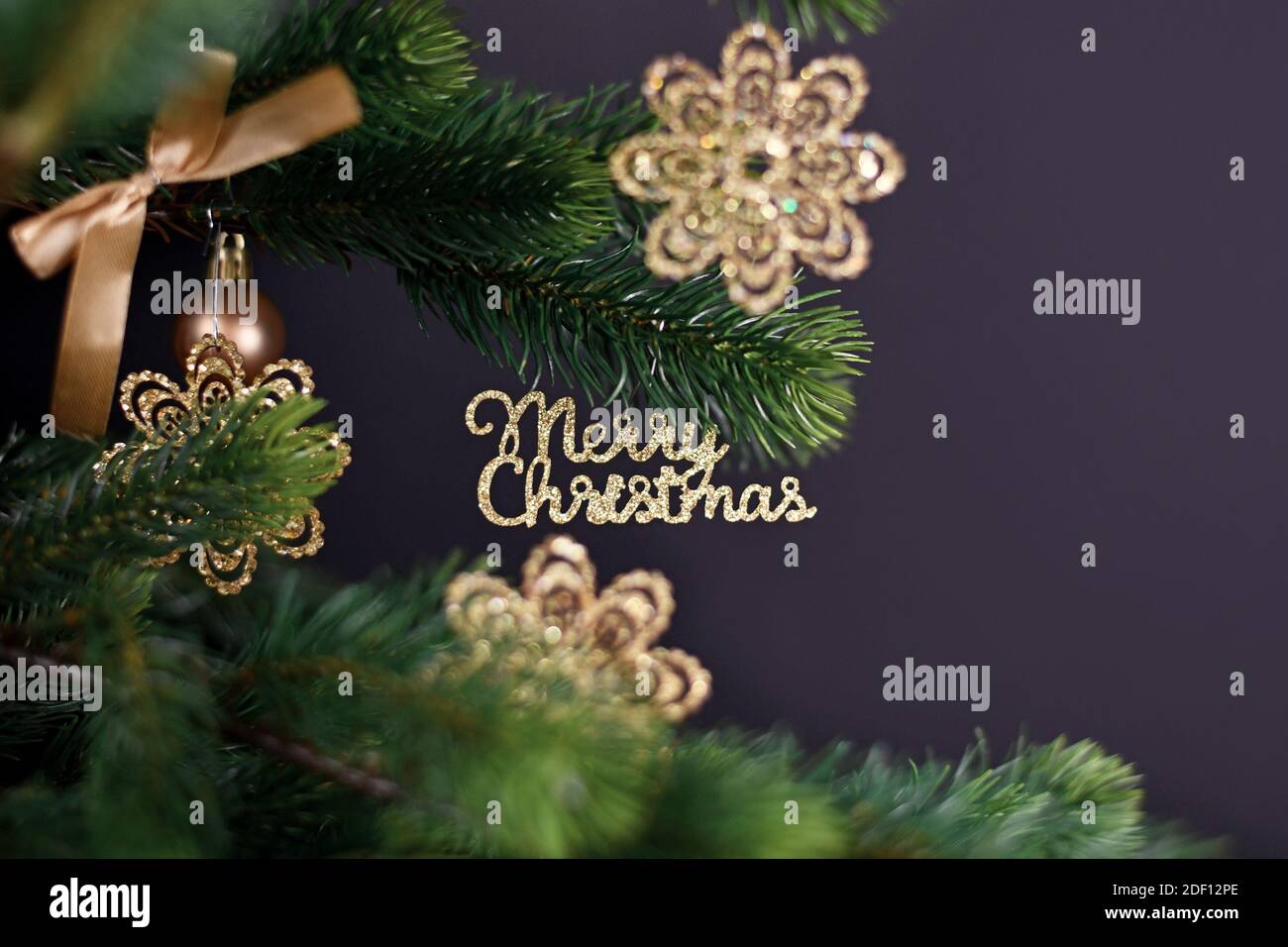 Goldener Weihnachtsbaum Ornament mit Text 'Merry Christmas' hängend aus Tannenzweig mit schneeflockenförmigen Kugeln auf dunklem Rücken Hintergrund Stockfoto