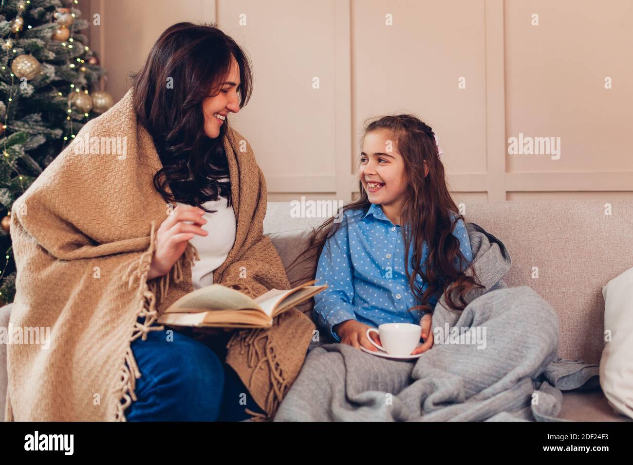 Junge Mutter und Tochter lesen Buch von Weihnachtsbaum im Wohnzimmer. Familie verbringt Zeit zusammen auf der Couch entspannen und Tee zu Hause trinken Stockfoto