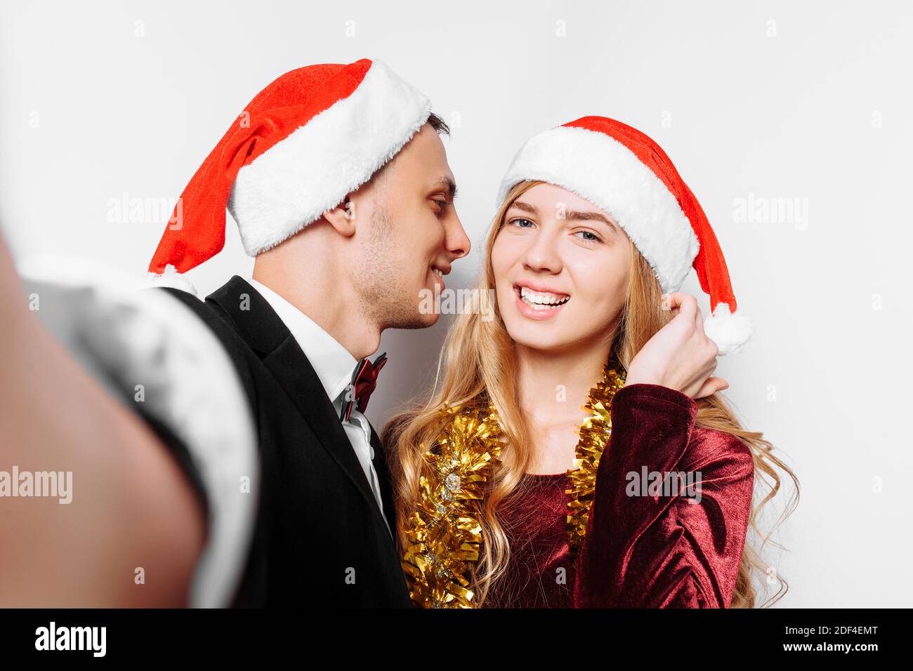 Glückliches Paar von Liebenden in Weihnachtsmannmützen, küsst der Kerl das Mädchen und macht ein Foto am Telefon, mit Weihnachtsgeschenken in ihren Händen. Auf einer weißen BA Stockfoto