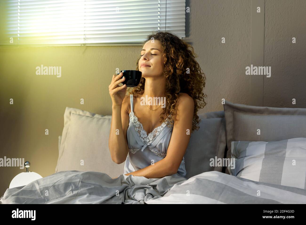 Schöne junge lockige Frau im Pyjama riecht das Aroma von Kaffee im Bett, sitzen und halten schwarze Tasse in einer Hand, genießen den Morgen mit Augen cl Stockfoto