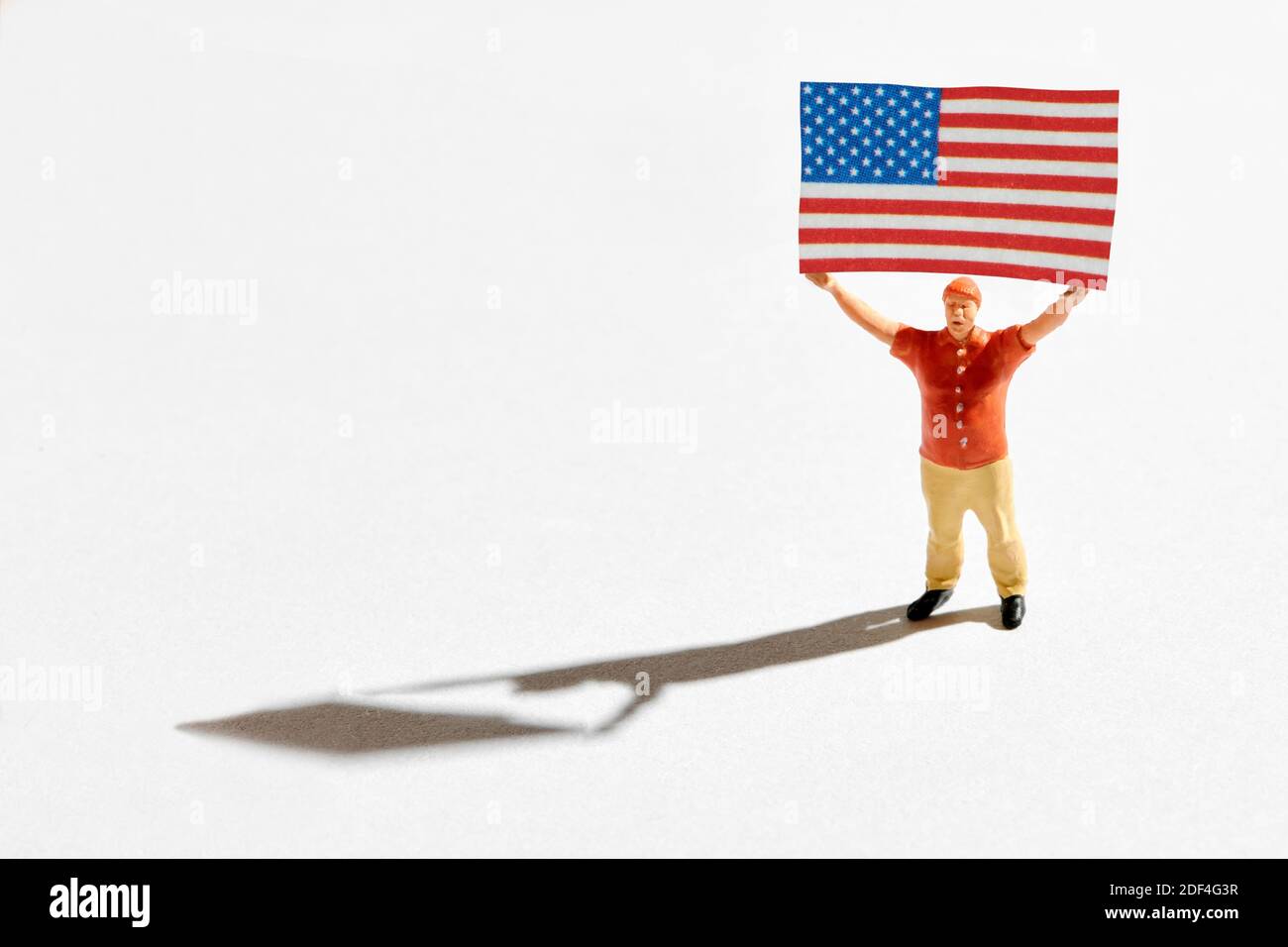 Politische Unterstützerfigur mit amerikanischer Flagge. Miniatur eines patriotischen Mannes in rotem Hemd, der die Flagge der USA in beiden Händen über den Kopf hält. Hoch und hoch Stockfoto