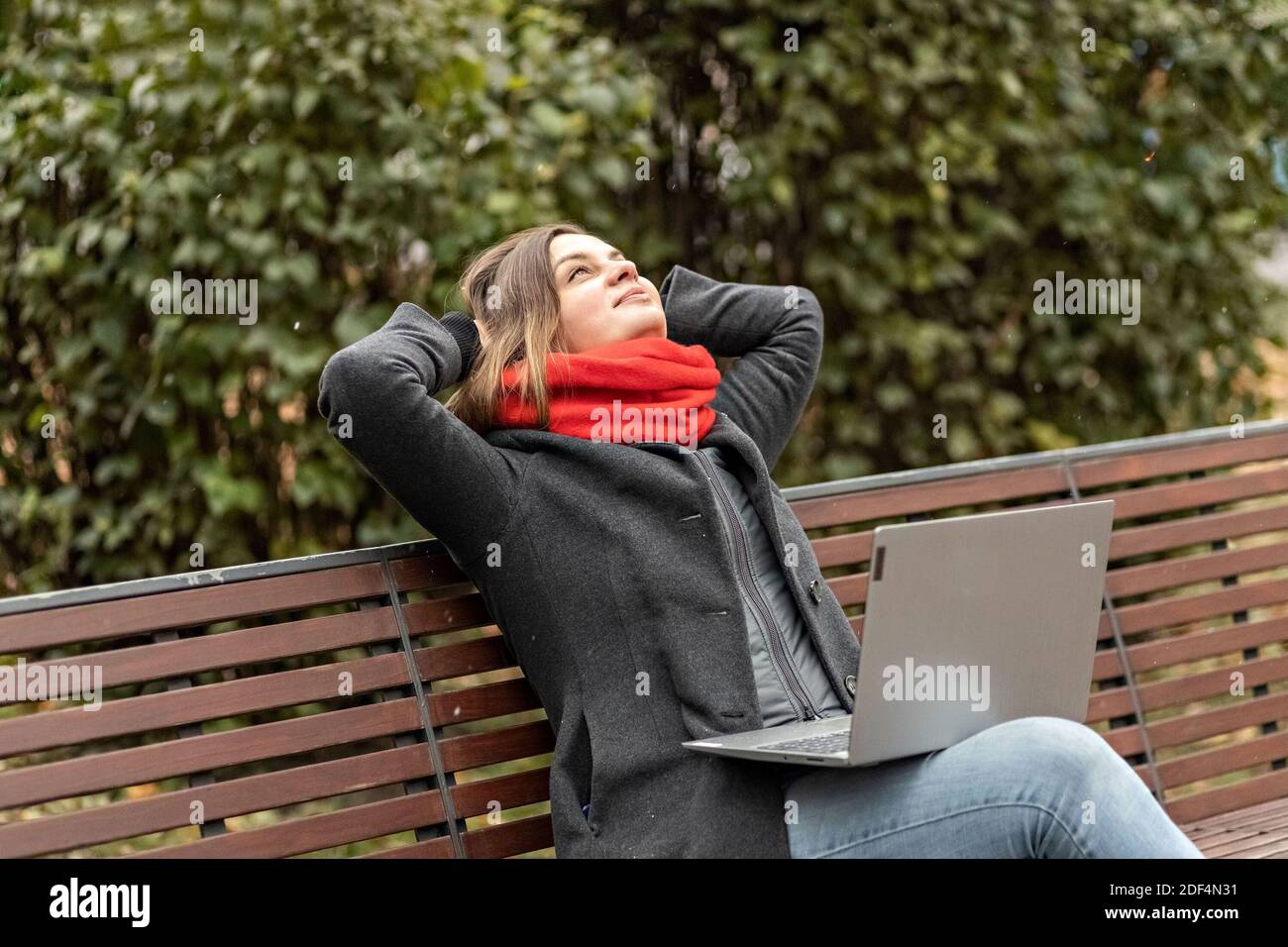 Eine junge Frau, mit den Händen hinter dem Kopf, fühlt Ruhe, ruht nach der Arbeit, sitzt auf einer Holzbank im Park ein Laptop auf den Knien. Pause während w Stockfoto