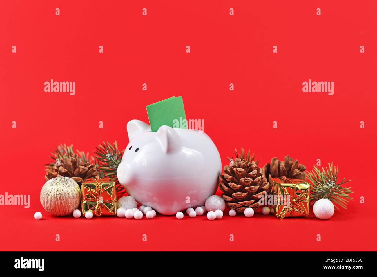 Weihnachtsarrangement mit Sparschwein mit grünem Gutschein umgeben von Saisonale Ornamente auf rotem Hintergrund Stockfoto