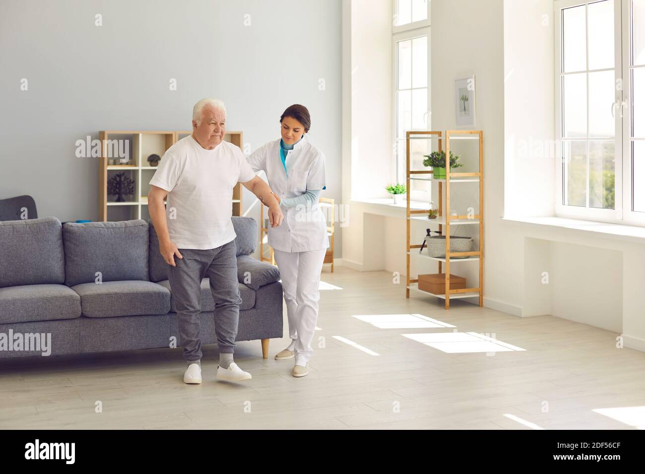 Eine freundliche Krankenschwester hilft einem älteren Patienten, mit der Hand durch den Raum in einem Pflegeheim zu gehen. Stockfoto