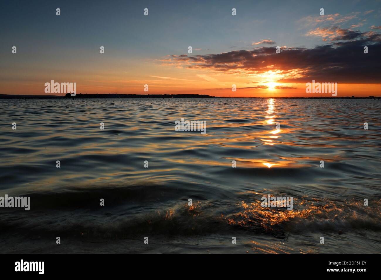 Die Sonne füllt den Horizont mit warmem, goldenem Licht, wenn sie durch dicke Wolken untergeht. Das Sonnenlicht funkelt auf den sanften, plätschernden Wellen eines ruhigen Meeres. Stockfoto