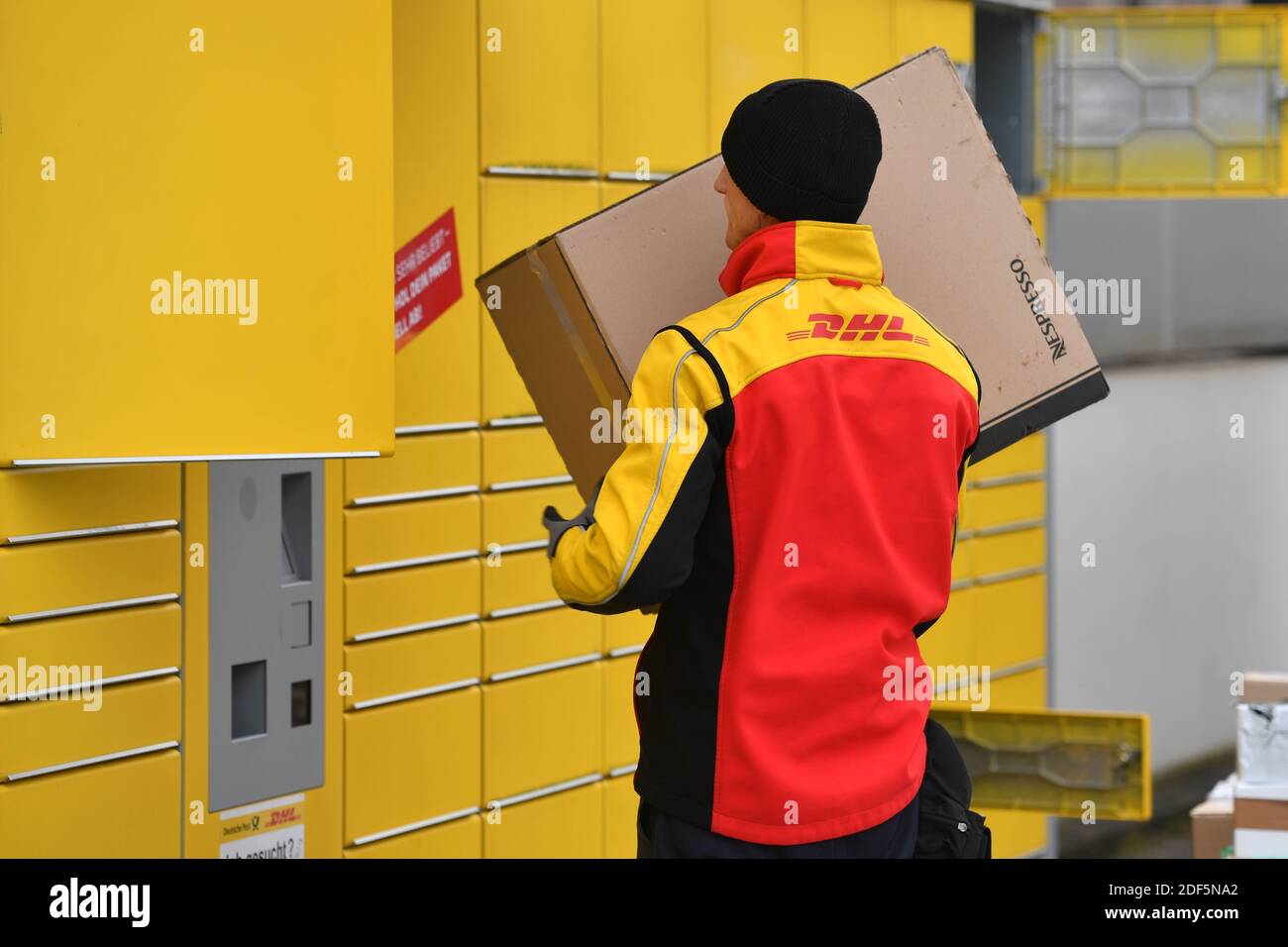 München, Deutschland. Dezember 2020. Paketvolumen und Online-Handel boomen in Zeiten der Corona-Virus-Pandemie. DHL Paketzusteller, Paketdienstleister, bei der Arbeit legt Paket in ein Fach einer Packstation. Entlädt seine Paket Auto, Postbus, DHL, Paket, Pakete, Paketdienst, liefern, liefern, entladen, Versand, Transporter, Handfahrzeug. Quelle: dpa/Alamy Live News Stockfoto