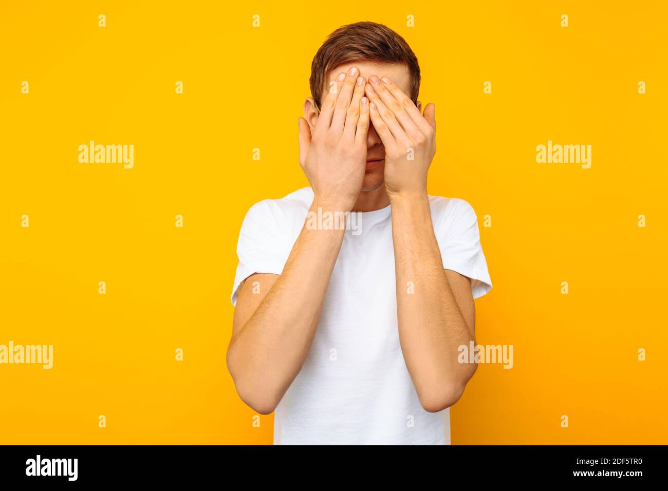Porträt eines verängstigten Mannes mit Brille, ein Mann in einem weißen T-Shirt, schloss seine Augen mit den Händen aus Angst, auf einem gelben Hintergrund Stockfoto