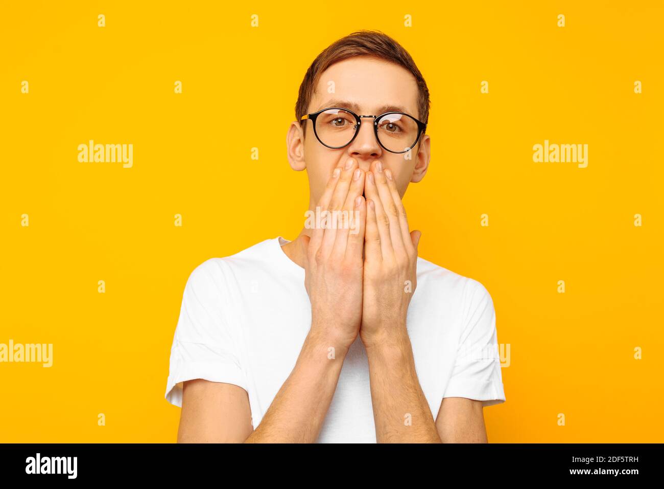 Porträt eines verängstigten Mannes mit Brille, eines Mannes, der in einem weißen T-Shirt gekleidet ist und vor Angst seinen Mund mit den Händen bedeckt, auf einem gelben Hintergrund Stockfoto