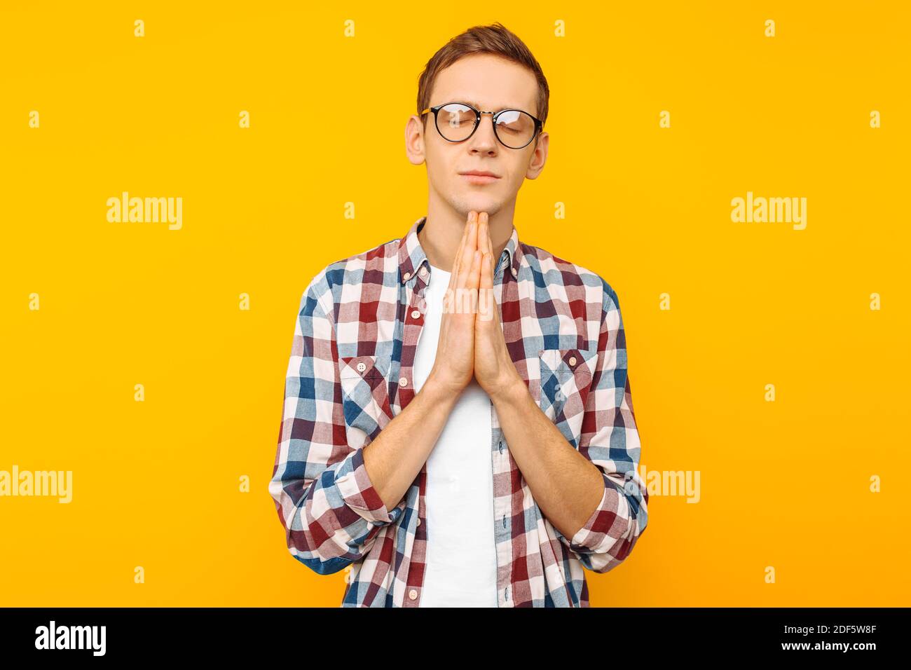 Porträt eines jungen Mannes mit Brille, ein Kerl in einem karierten Hemd, der seine Hände im Gebet oder Meditation faltete, sah entspannt und ruhig aus, träumte und war w Stockfoto