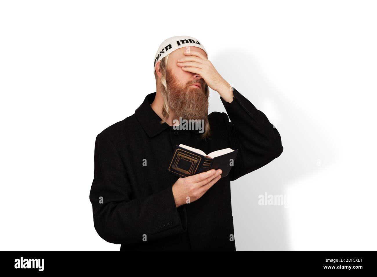 Frommer jüdischer Mann, der Siddur in seiner Hand hält, die Augen mit der Hand bedeckt, die auf weißem Hintergrund isoliert ist. Bärtiger Jude chassid mit Seitenschwänzen in weißem Kippach Stockfoto