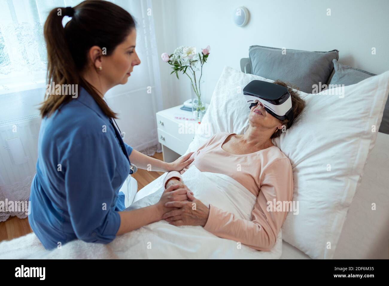 Ältere Patientin, die sich im Krankenhausbett entspannt, mit Virtual Reality-Headset. Startseite Krankenschwester sitzen auf dem Bett Blick auf ältere Frau bekommen VR-Behandlung. Stockfoto