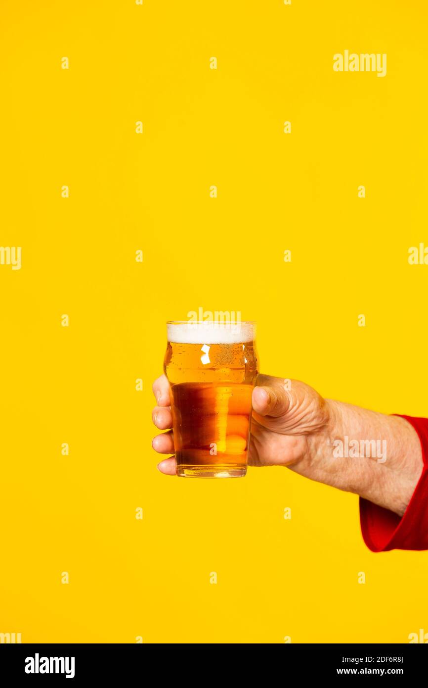 Hand einer älteren Frau, die ein Glas eines halben Pint Bier vor einem gelben Hintergrund hält Stockfoto