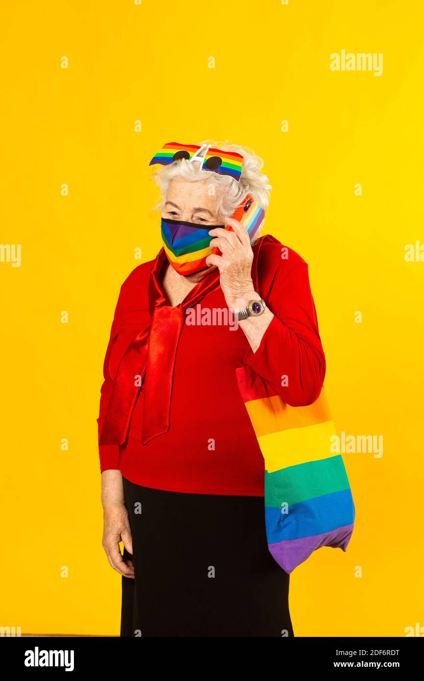 Studio Portrait einer älteren Frau in einem roten Hemd, Regenbogensonnenbrille, Regenbogenfarben Gesichtsmaske und einer Tasche, einen Anruf mit ihrem Handy, Stockfoto