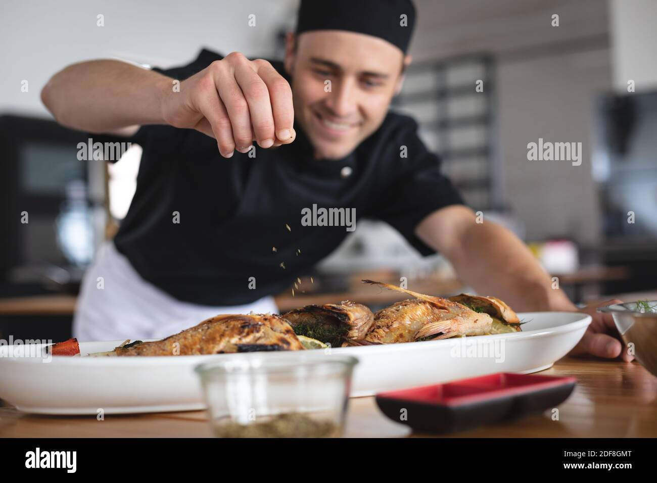 Männlicher kaukasischer Koch, der Salz über geröstete Fischgerichte zugibt Teller in der Restaurantküche Stockfoto