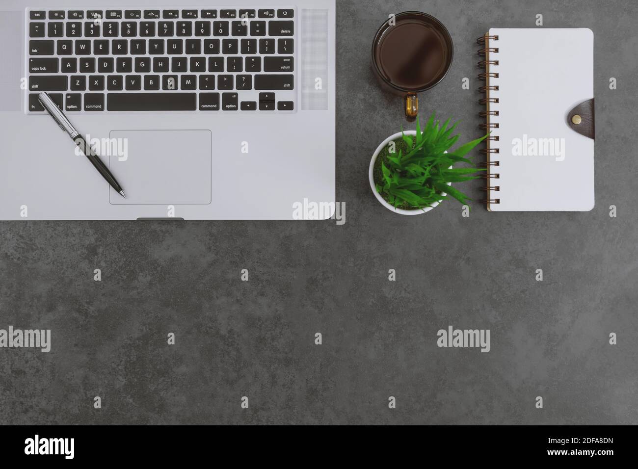 Blick von oben auf den Schreibtisch mit Laptop, Notebook, Stift und Topfpflanze auf dunkelgrauem Hintergrund. Stockfoto