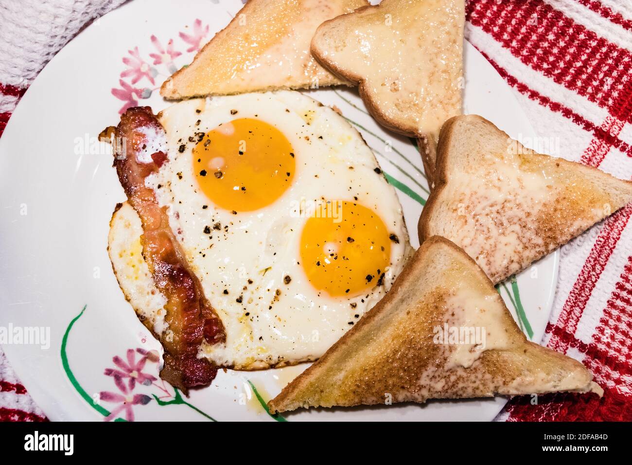 Nahaufnahme der sonnigen Seite up Eier mit Speck und Brot auf der Seite. Teller mit Blumen Design. Hintergrund - weiße und rote Tischdecke Stockfoto