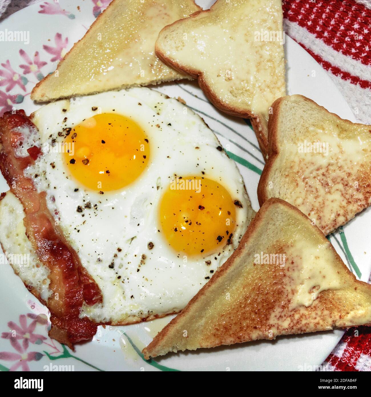 Nahaufnahme der sonnigen Seite up Eier mit Speck und Brot auf der Seite. Teller mit Blumen Design. Hintergrund - weiße und rote Tischdecke Stockfoto