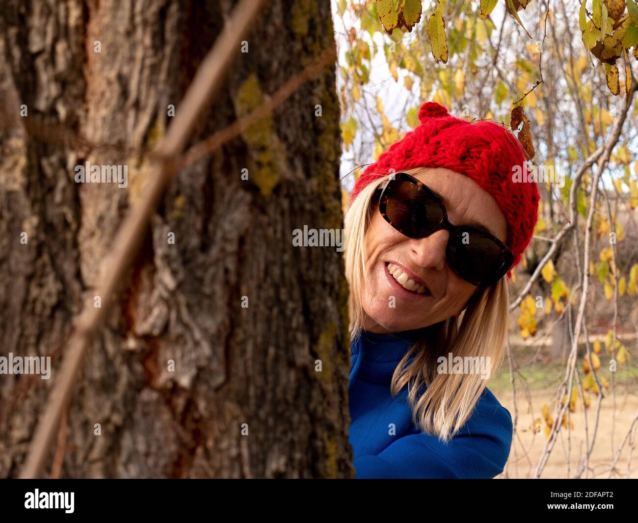 Ein flacher Fokus einer fröhlichen blonden Frau mittleren Alters Blick hinter einem Baum in einen Park Stockfoto