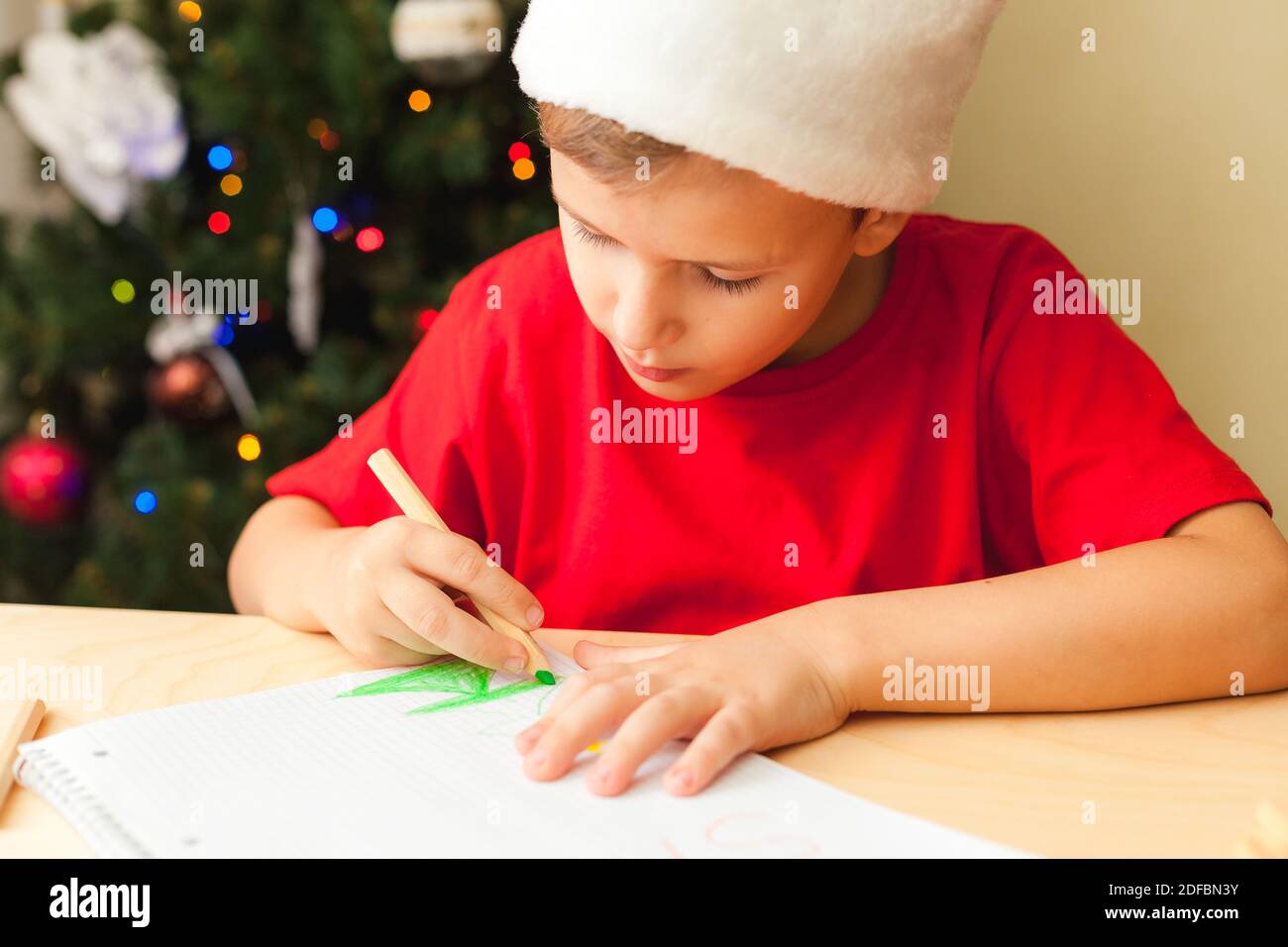 Nettes Kind Junge trägt roten Hut schreiben Brief an Santa sitzen am Schreibtisch, Weihnachtsbaum auf dem Hintergrund Stockfoto