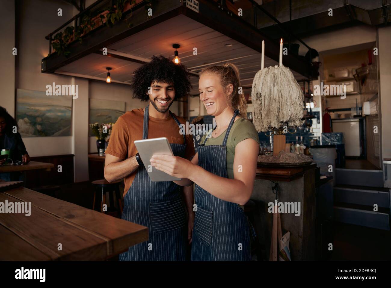 Glückliche männliche und weibliche Kollegen tragen Schürze und lachen, während Video in einem digitalen Tablet im Café ansehen Stockfoto