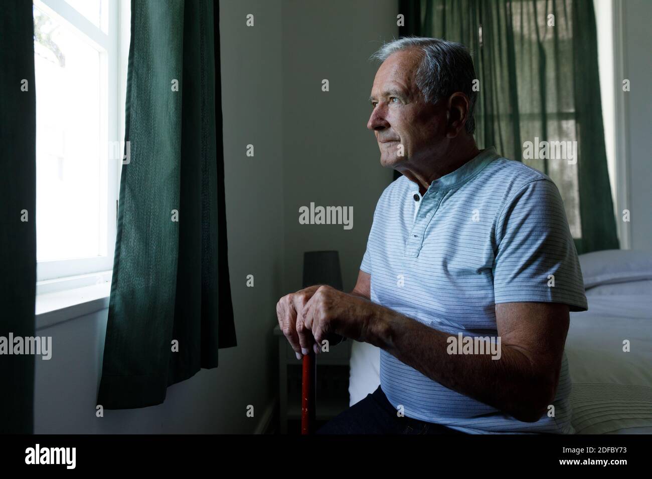 Älterer kaukasischer Mann, der einen Spazierstock hält und aus dem Fenster schaut Zu Hause Stockfoto