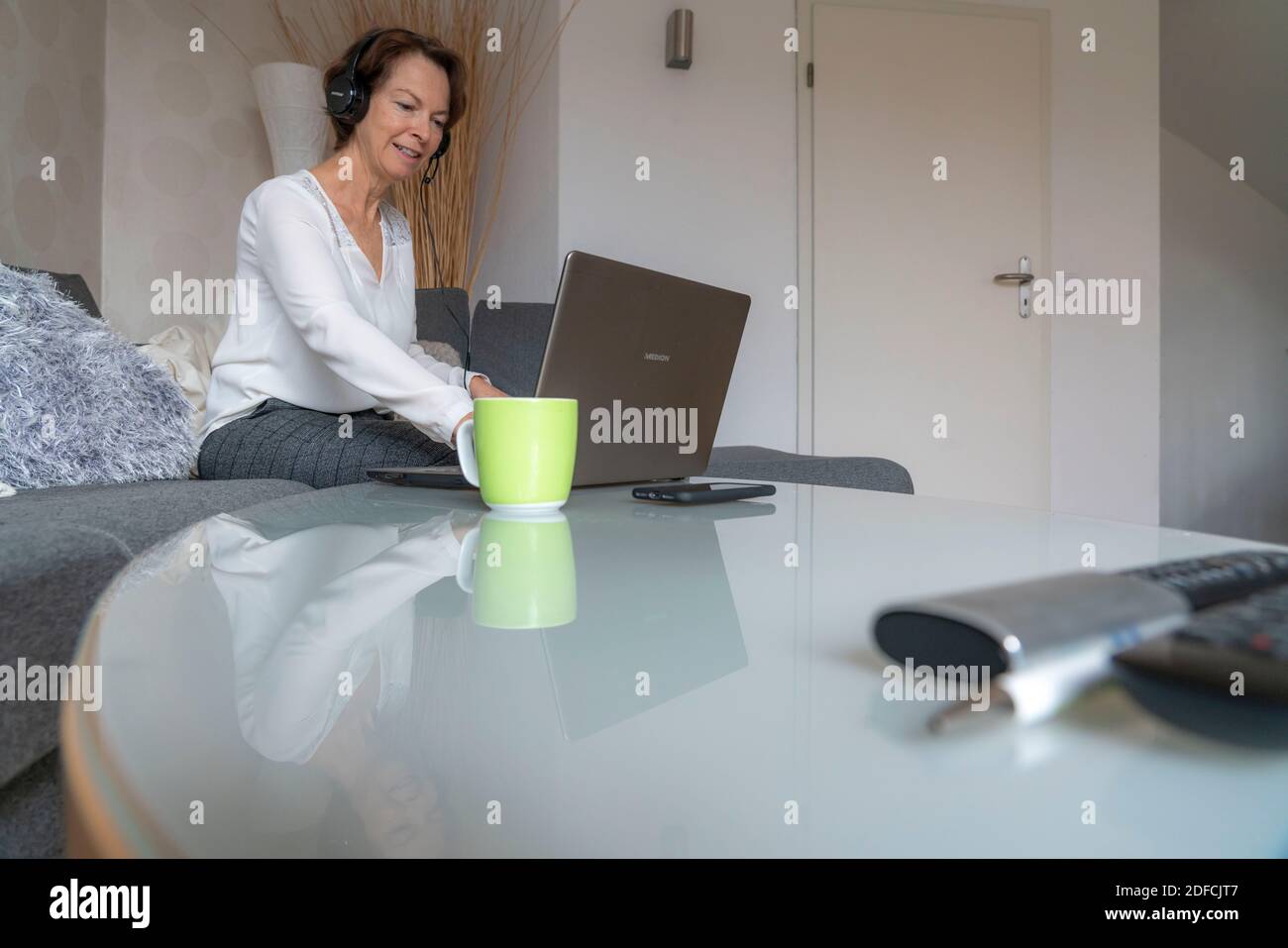 Frau, Mitte 50, arbeitet von zu Hause, mit Laptop und kommuniziert über Headset mit Kollegen, Home Office, auf dem Sofa, Stockfoto