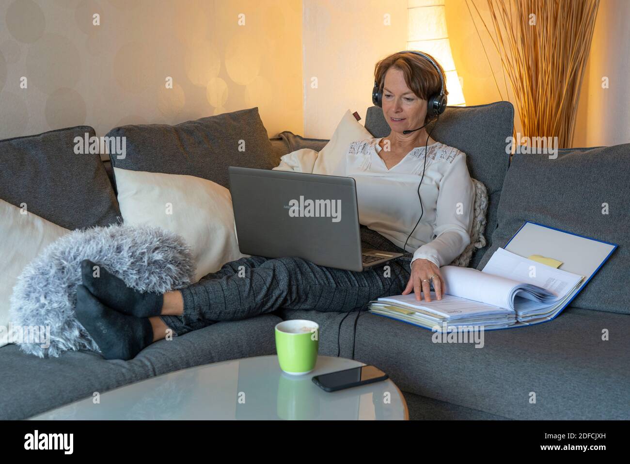 Frau, Mitte 50, arbeitet von zu Hause, mit Laptop und kommuniziert über Headset mit Kollegen, Home Office, auf dem Sofa, Stockfoto