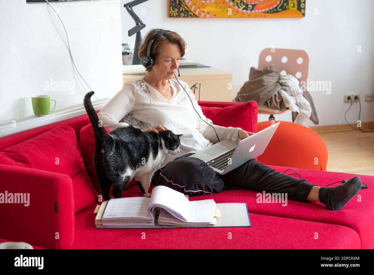 Frau, Mitte 50, arbeitet von zu Hause, mit Laptop und kommuniziert über Headset mit Kollegen, Home Office, auf dem Sofa, Stockfoto