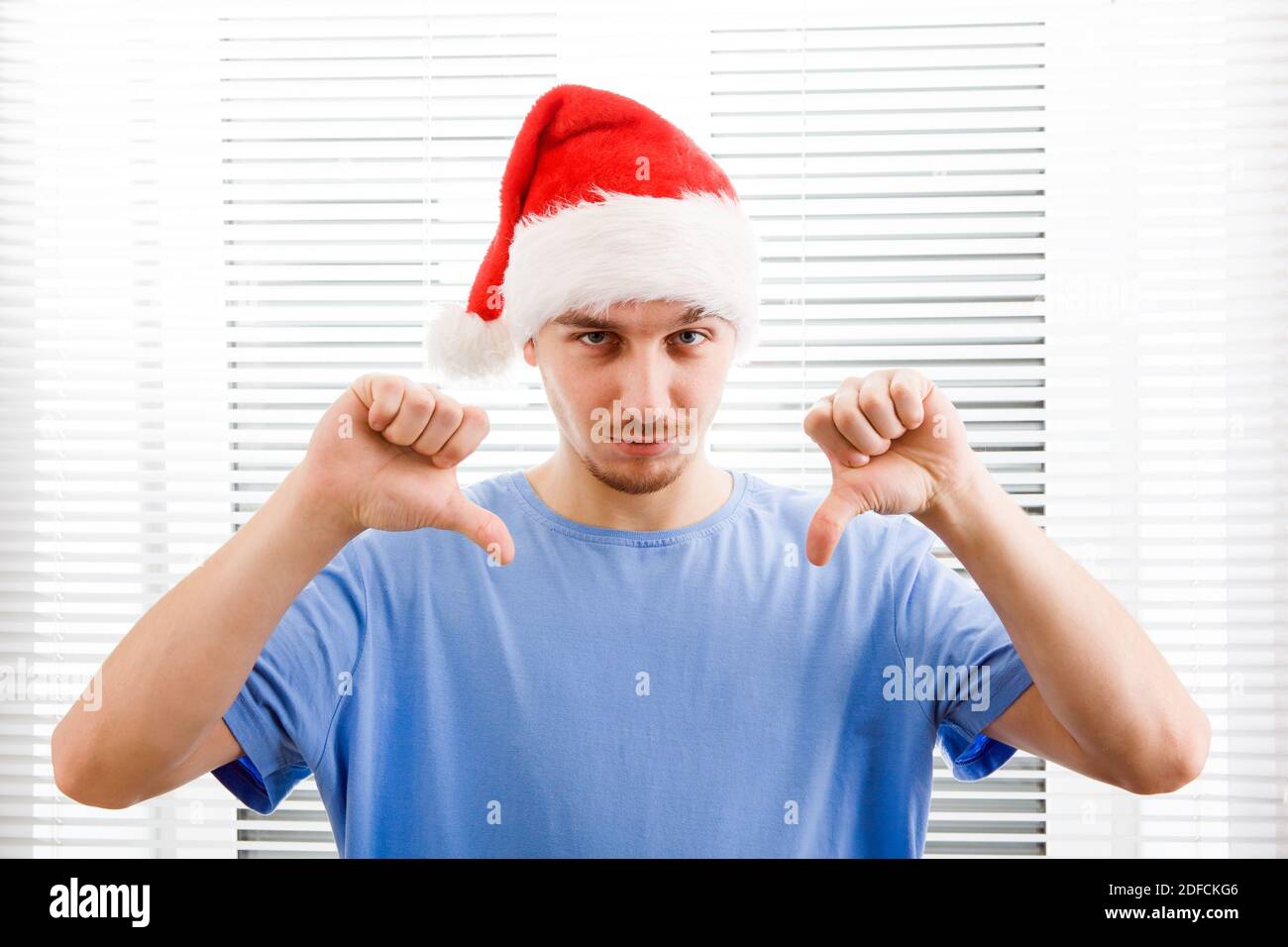 Junger Mann in Santa hat machen Daumen nach unten auf die Jalousie Hintergrund Stockfoto