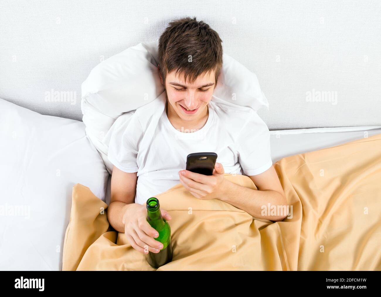 Fröhlicher junger Mann mit einem Telefon und einer Bierflasche Im Bett Stockfoto